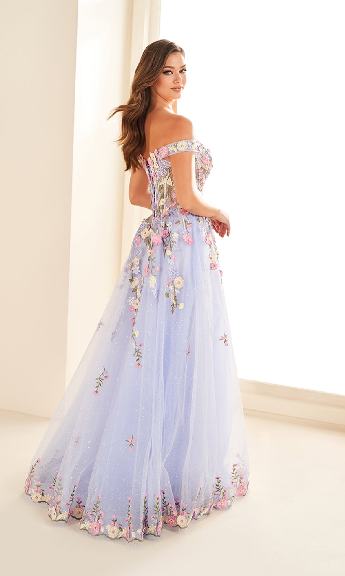 Woman wearing the Ellie Wilde EW36215 long prom dress, featuring off-the-shoulder floral embroidery, stands in a softly lit room and glances over her shoulder.